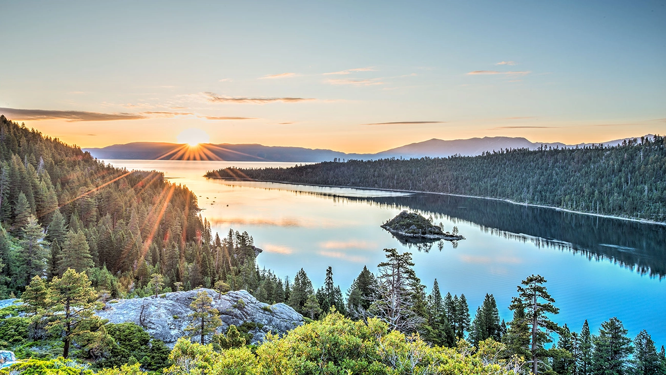 The sun peaks over a mountain illuminating the nearby trees and calm lake