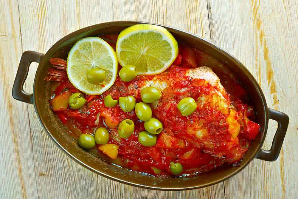 Veracruz-Style Red Snapper in a pan garnished with red sauce, green olives, and lemon slices.