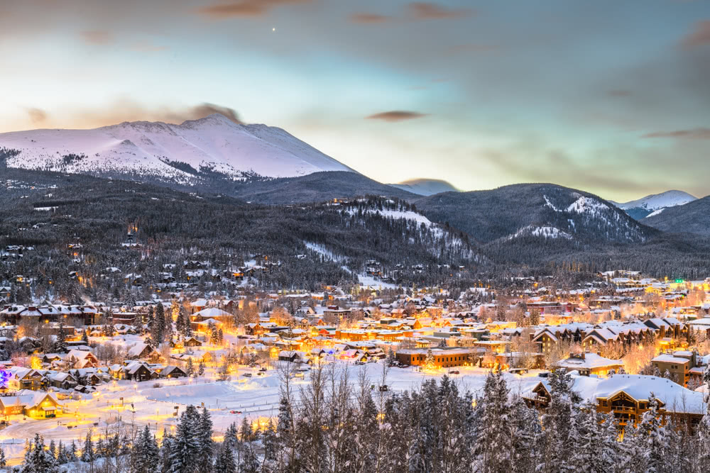 Breckenridge, Colorado city view at dusk with golden lights glowing on the snow and snowcapped mountains in the background.