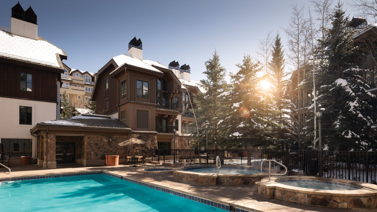 Two steaming hot tubs sit poolside as the sun shines through the trees