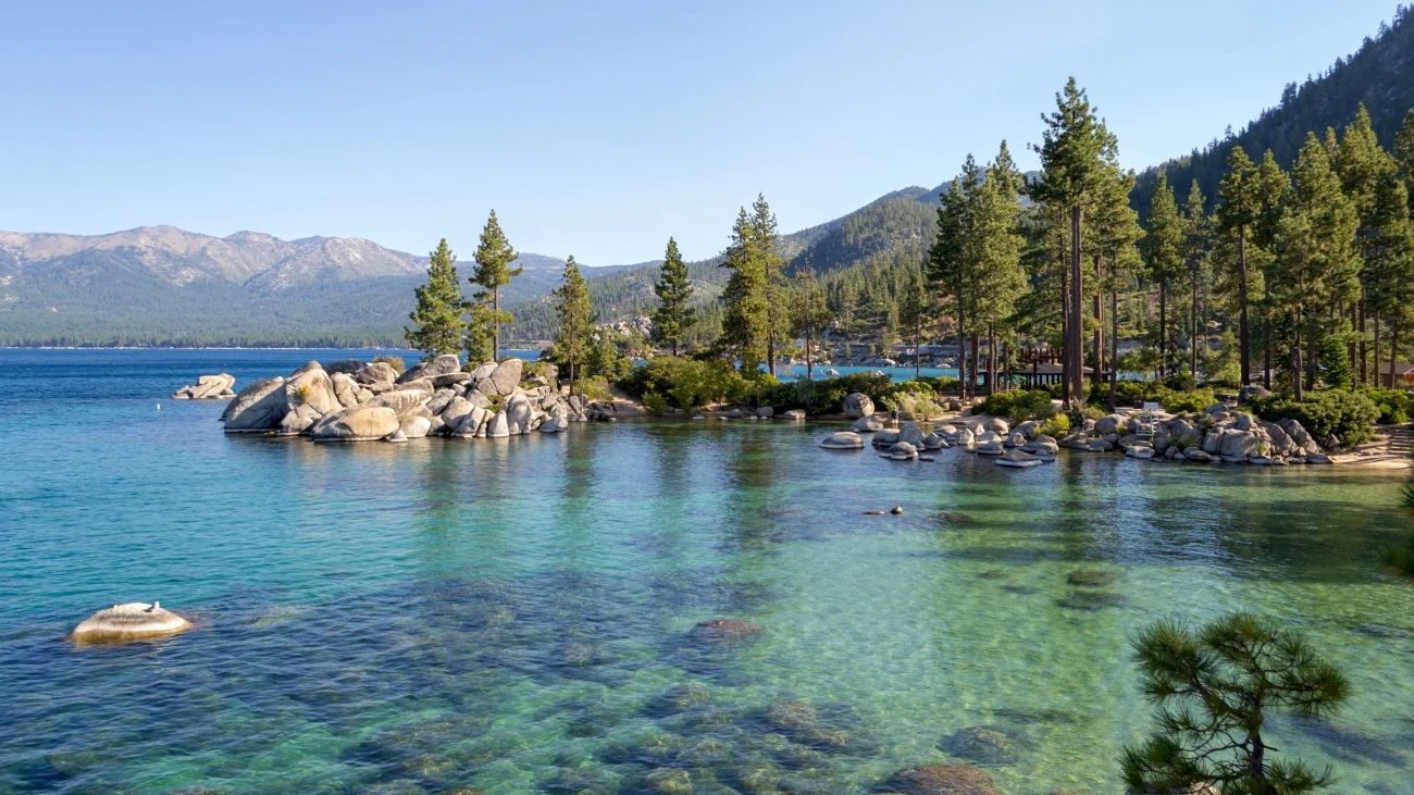 A crystal-clear shimmering lake with pine trees lining the rocky shore and mountains in the background