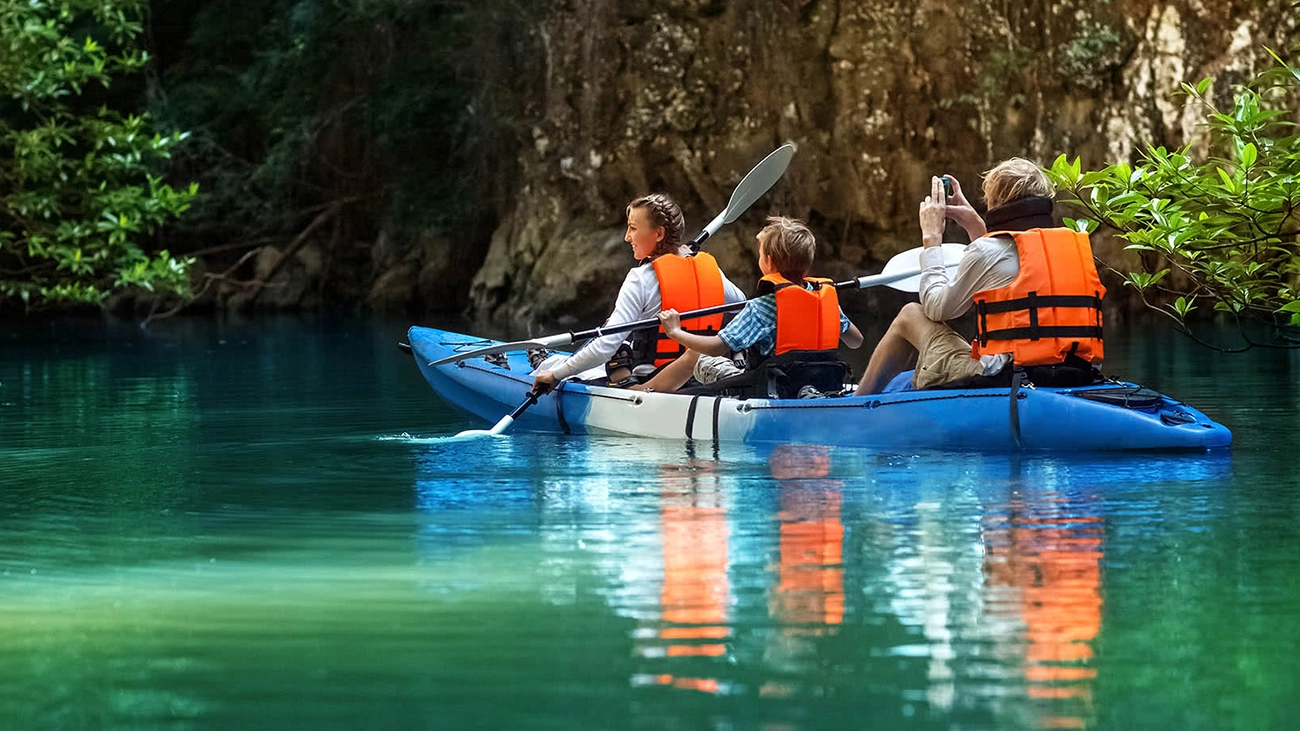 People in a three-person kayak in the water surrounded by with lush foliage