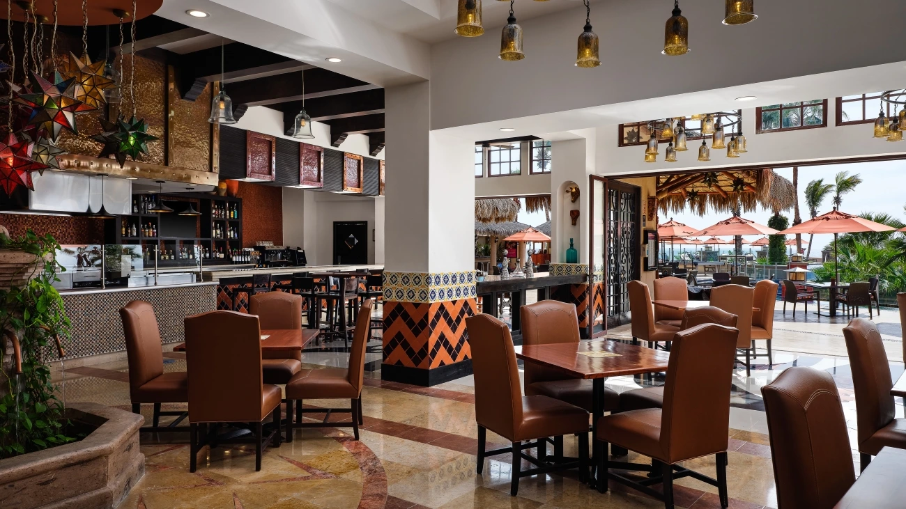 Restaurant and bar with dining tables near a patio with palm trees, tables, chairs, and umbrellas