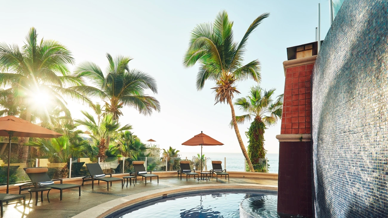 Poolside view with lounge chairs and umbrellas surrounded by palm trees by the ocean