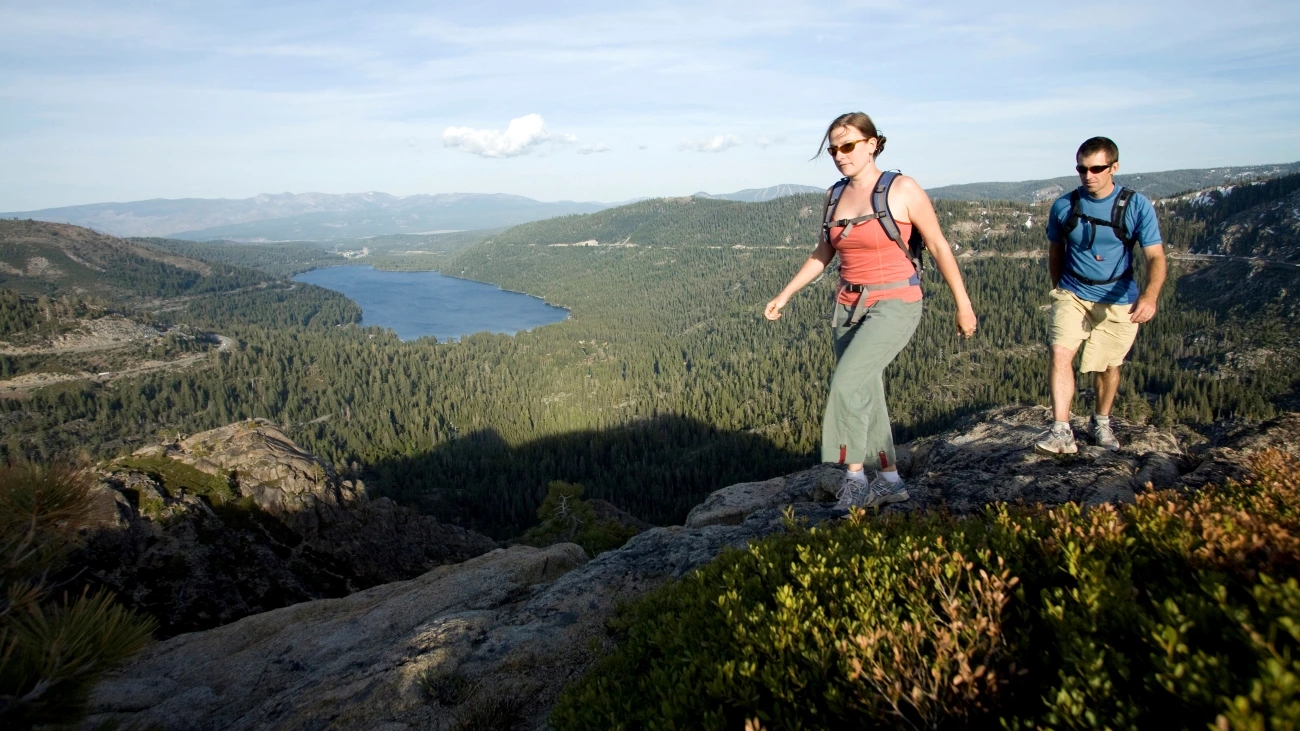 Couple hiking along scenic hilly terrain, with green forest and sparkling lake in the background